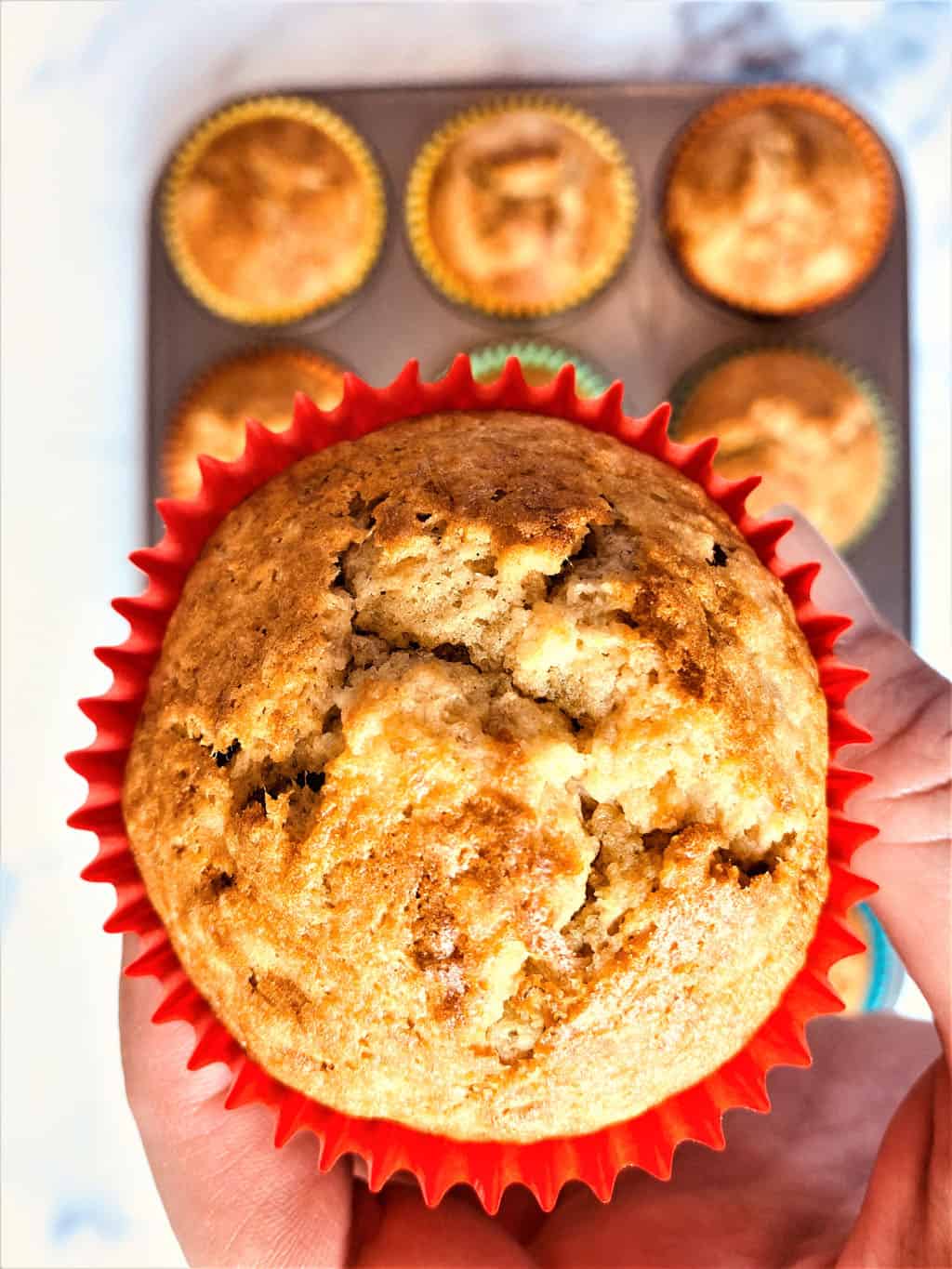 close up of banana muffin from above in a red muffin case with the remaining muffins below in baking tray