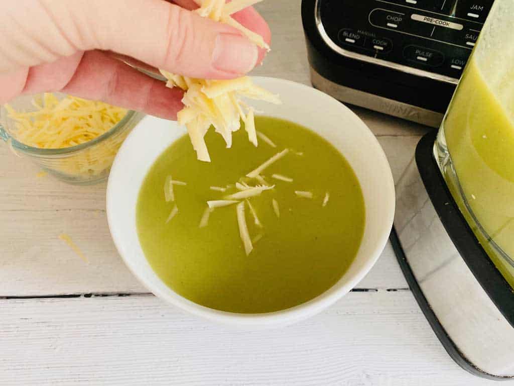 sprinkling grated cheddar cheese into hot broccoli soup next to Ninja soup maker