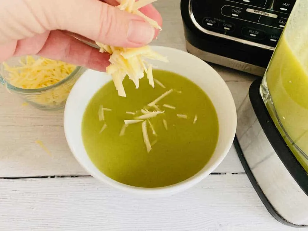 sprinkling grated cheddar cheese into hot broccoli soup next to Ninja soup maker