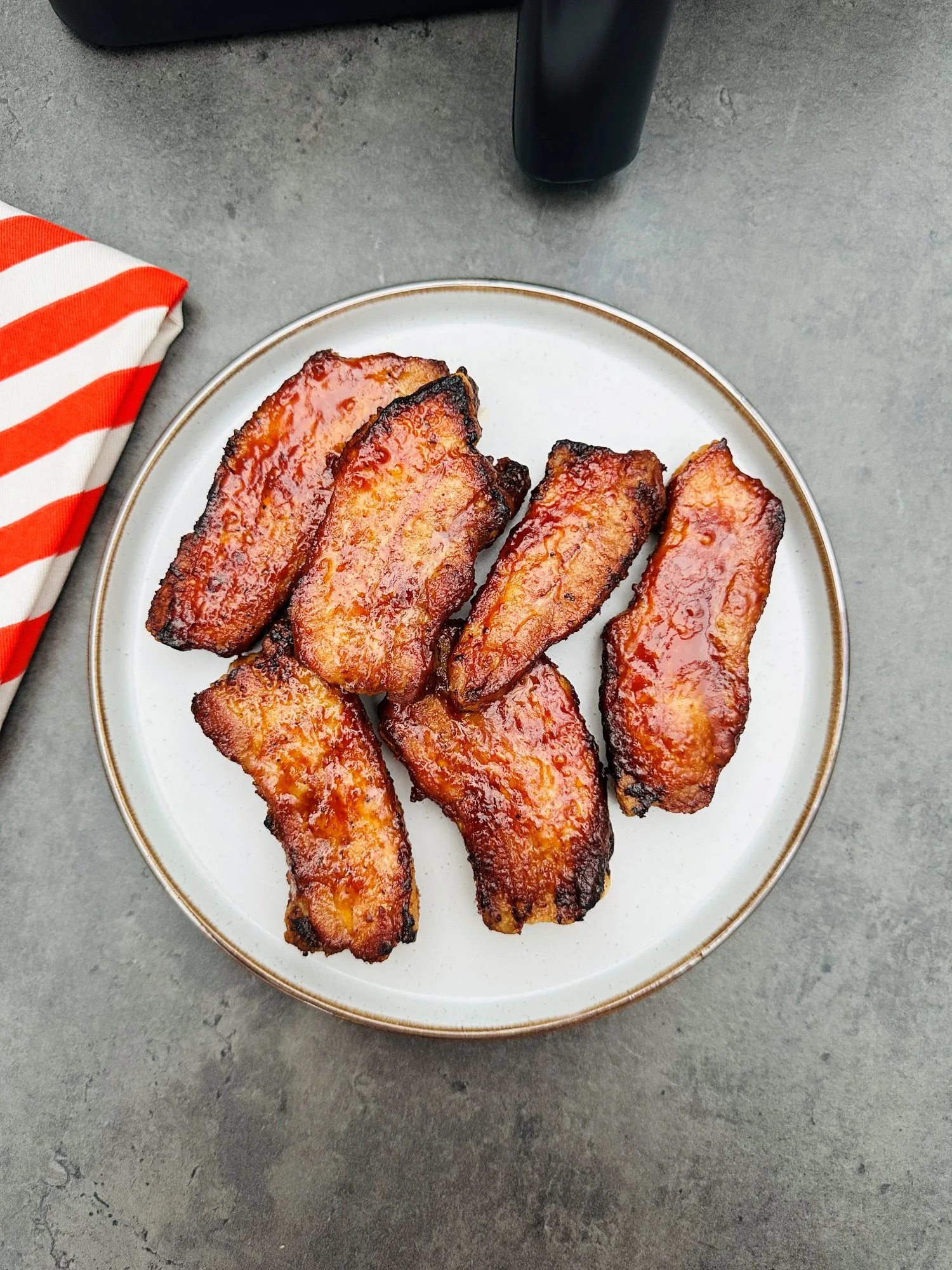 BBQ pork belly slices on a plate next to a red and white striped tea towel and an air fryer