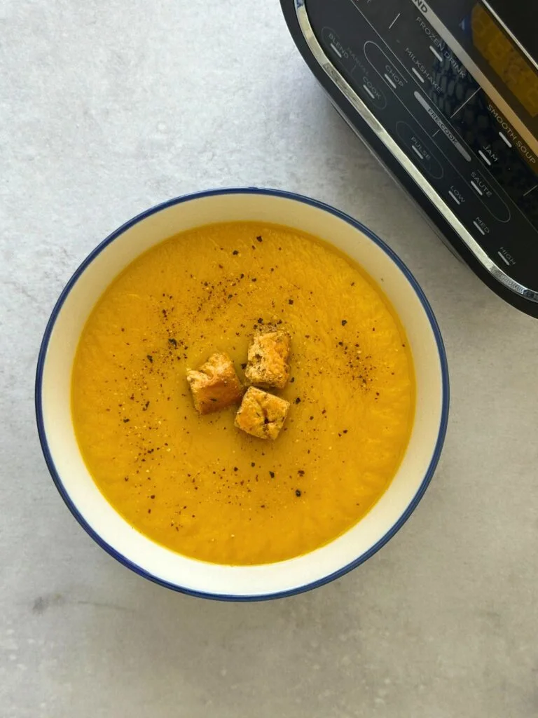 carrot and parsnip soup in a bowl with croutons and a pepper seasoning next to a Ninja Soup Maker