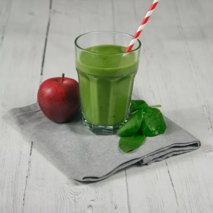 green smoothie in a glass with a red and white striped straw. Red apple and fresh spinach leaves next to glass on a grey tea towel.