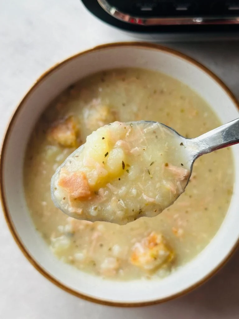 a spoon holding some chunky ham and potato soup above a bowl of soup