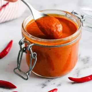 homemade Harissa Paste in a glass jar with a spoon dipped in surrounded by red chillies