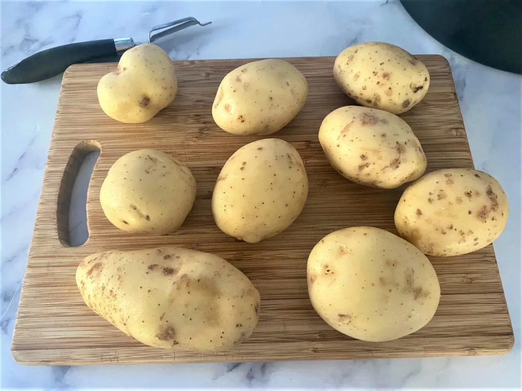 Maris Piper potatoes on chopping board with vegetable peeler ready to be peeled for roasting.