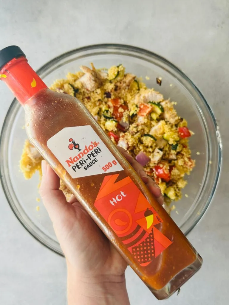 Nando's Peri Peri Sauce (Hot) bottle held over a glass bowl of couscous with vegetables and chicken