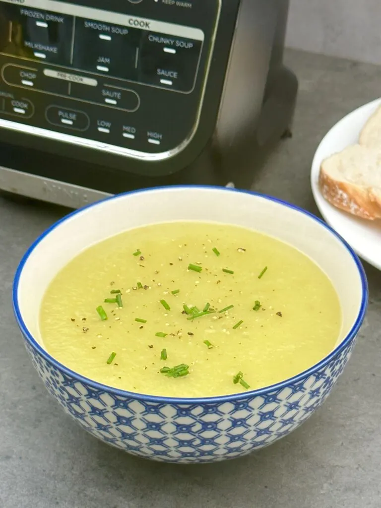 leek and potato soup in a bowl with chives and ground pepper on top. There is a slice of white bread on a white plate and a Ninja soup maker in the background