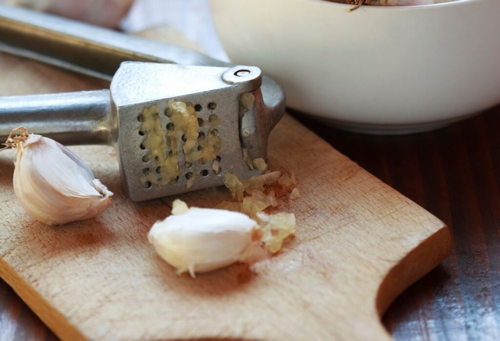 garlic cloves being crushed with a garlic press on wooden chopping board
