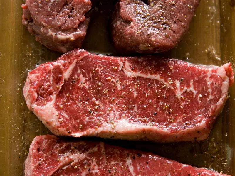 raw steak on chopping board being seasoned with salt and pepper