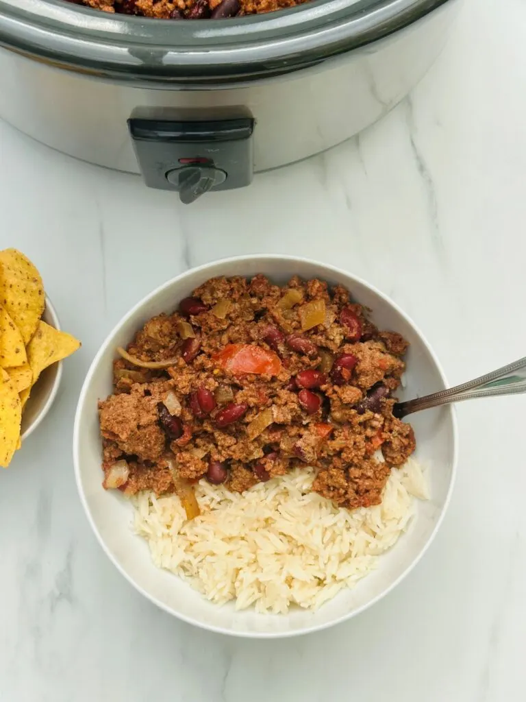 chilli con carne with rice in a bowl next to slow cooker and bowl of tortilla chips
