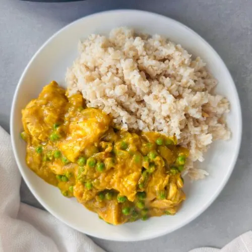 Chinese chicken curry served on a white plate next to some cooked rice with part of a slow cooker showing in the corner