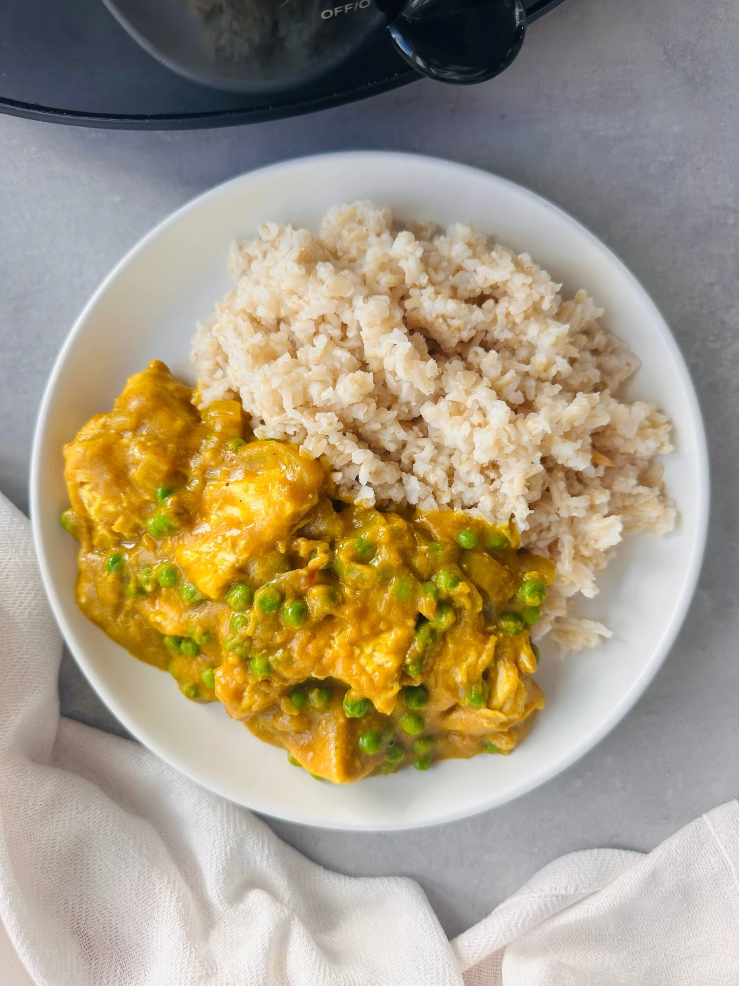 Chinese chicken curry served on a white plate next to some cooked rice with part of a slow cooker showing in the corner