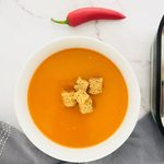 red pepper and chilli soup in a bowl with croutons on top and a fresh red chilli next to it. also in the photo is a grey tea towel and a small part of the ninja soup maker