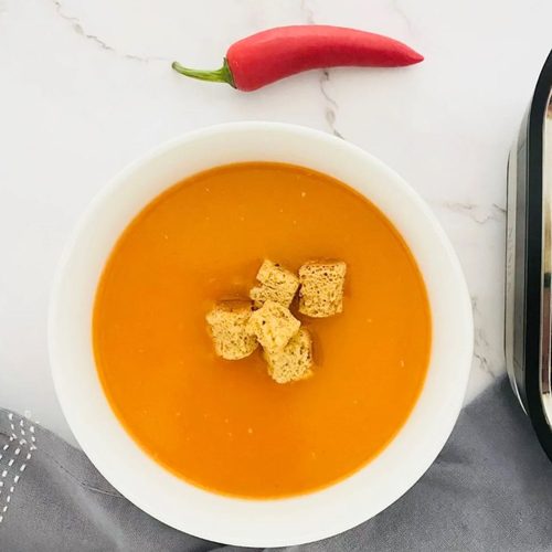 red pepper and chilli soup in a bowl with croutons on top and a fresh red chilli next to it. also in the photo is a grey tea towel and a small part of the ninja soup maker