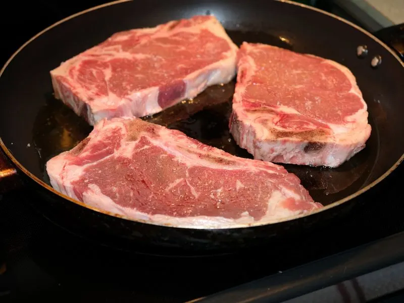 three raw steaks in a pan frying