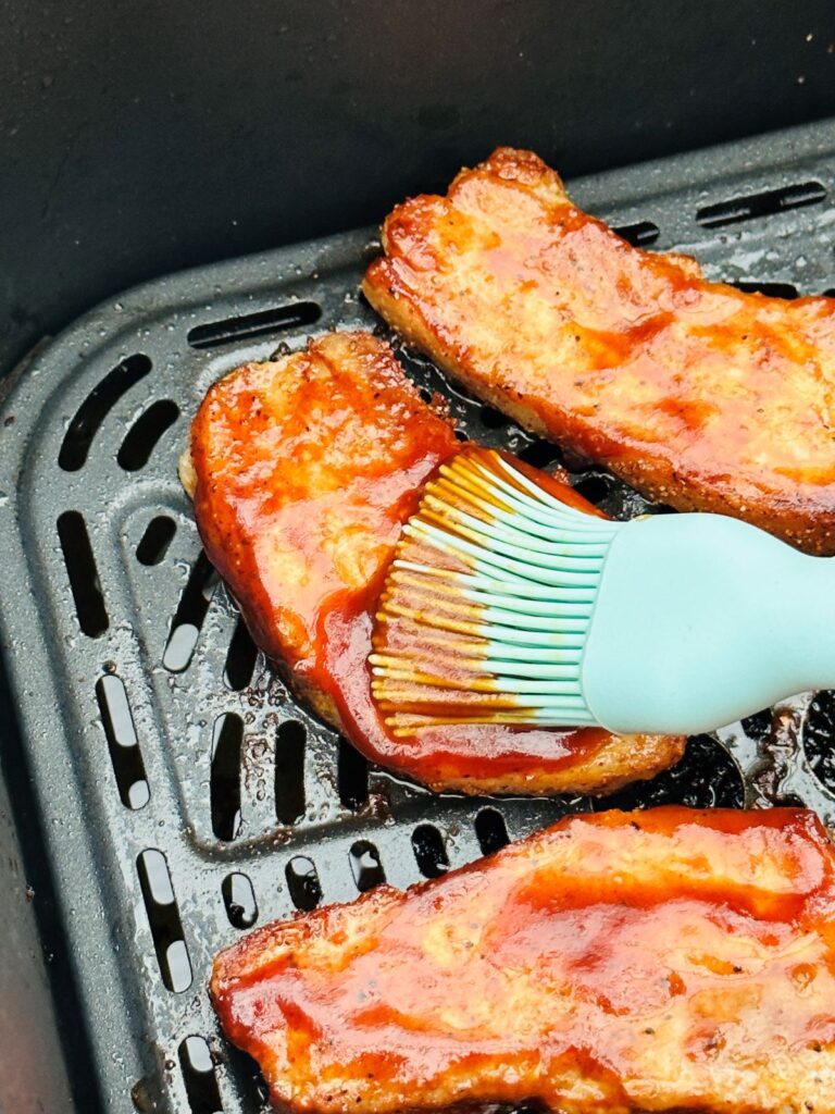 brushing BBQ sauce over pork belly slices in air fryer basket