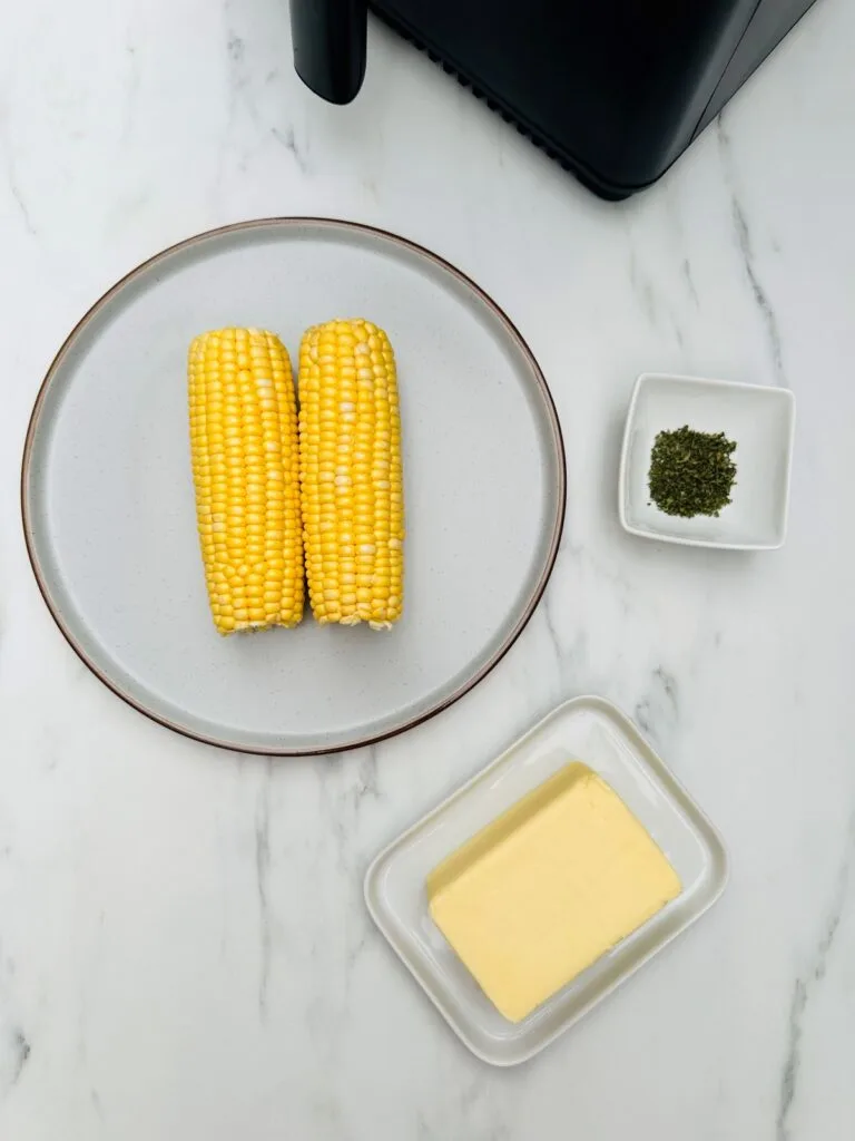 two uncooked corn on the cobs on a plate, butter, parsley, air fryer on a white background 