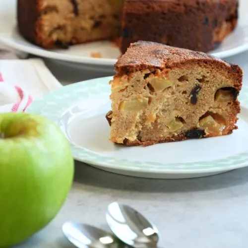 slice of apple and cinnamon cake on a plate with two teaspoons and an apple in front with the remaining cake in the background
