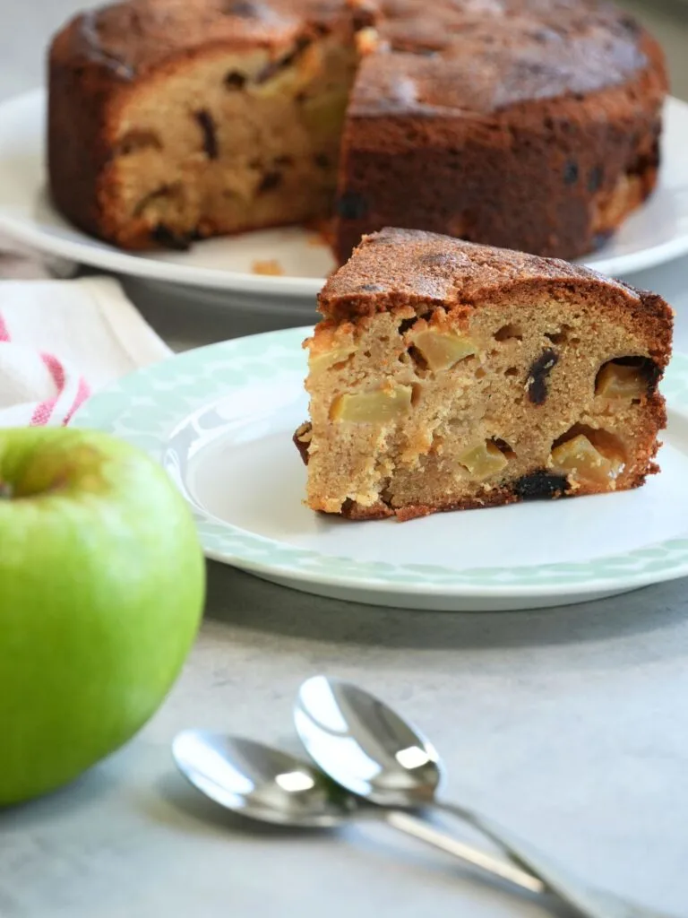 slice of apple and cinnamon cake on a plate with two teaspoons and an apple in front with the remaining cake in the background