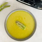 asparagus soup in a bowl with two tips for garnish. the soup bowl is positioned next to the Ninja soup maker and some fresh raw asparagus.