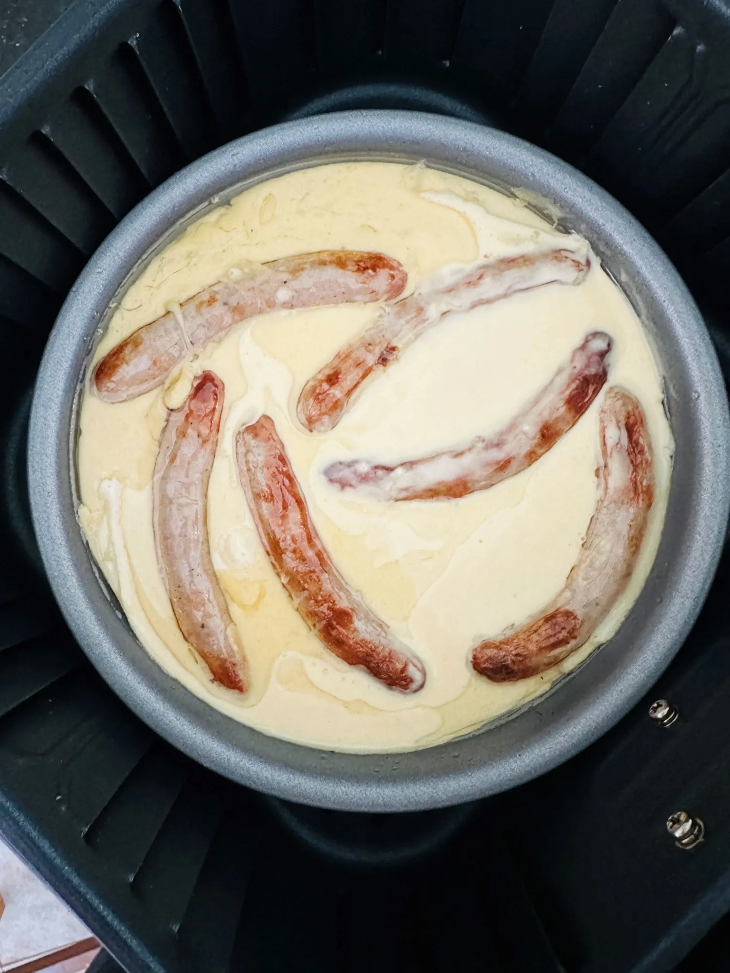 batter poured over the browned sausages in a 20 cm sandwich tin in an air fryer basket ready to be air fried