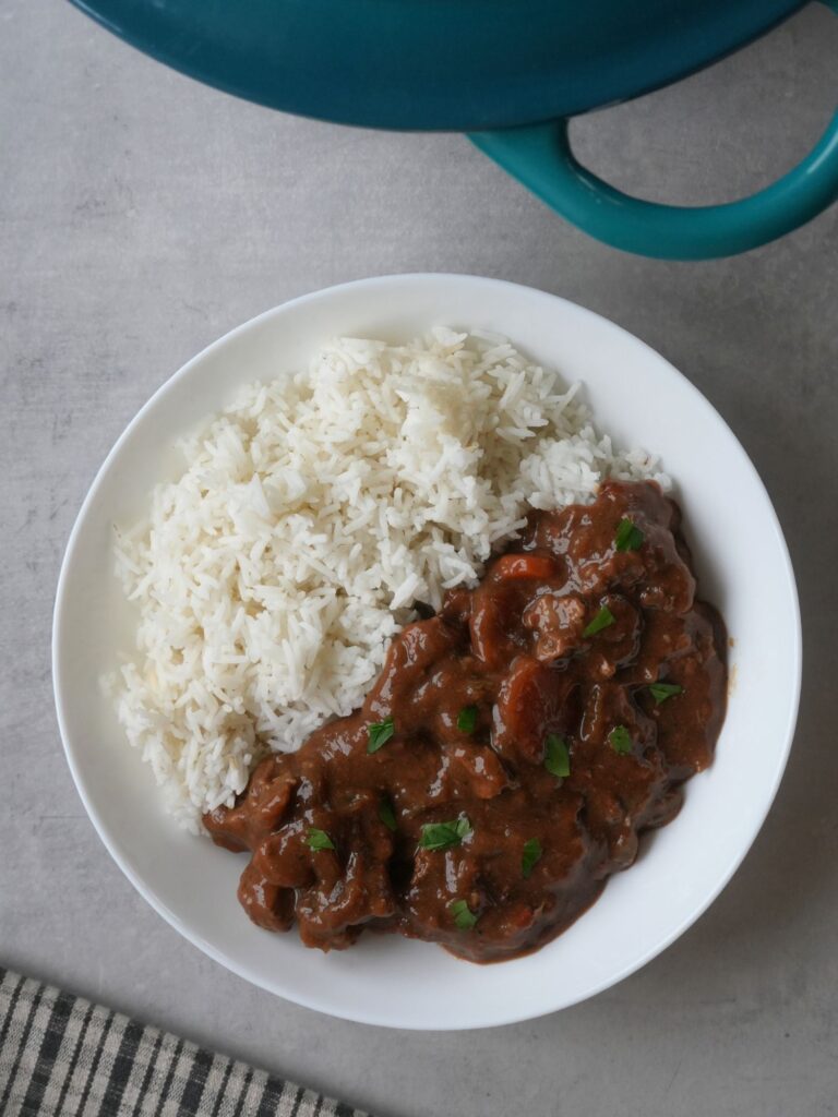 beef and ale stew next to rice on a plate with part of a casserole dish showing at the edge