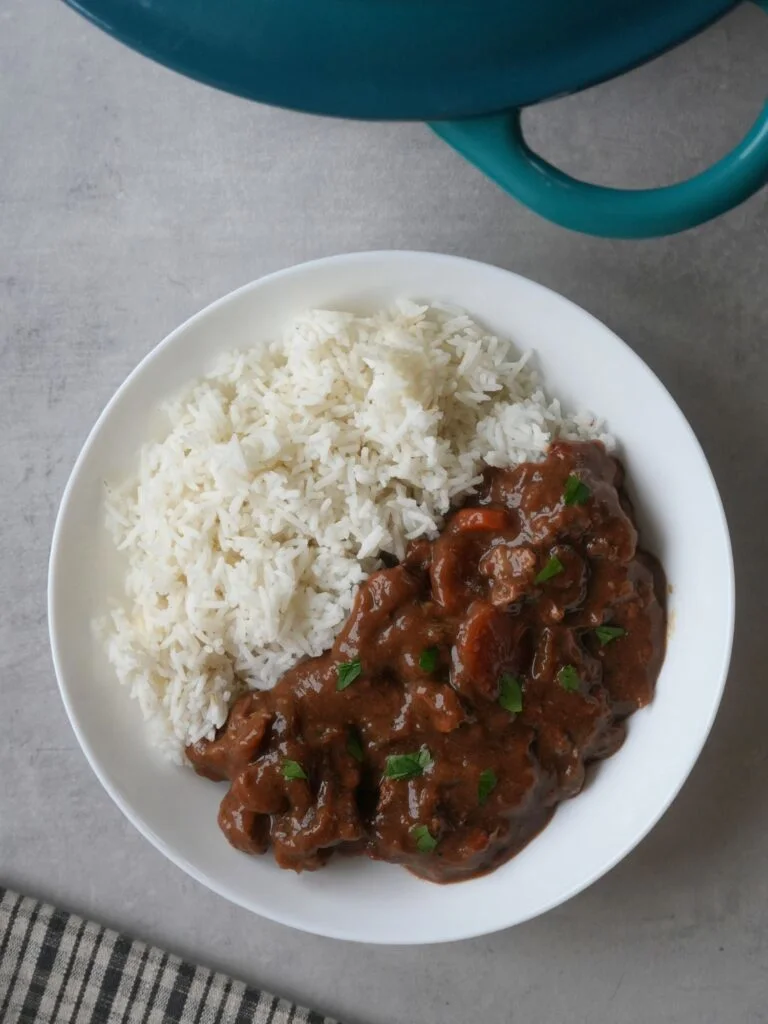 beef and ale stew next to rice on a plate with part of a casserole dish showing at the edge