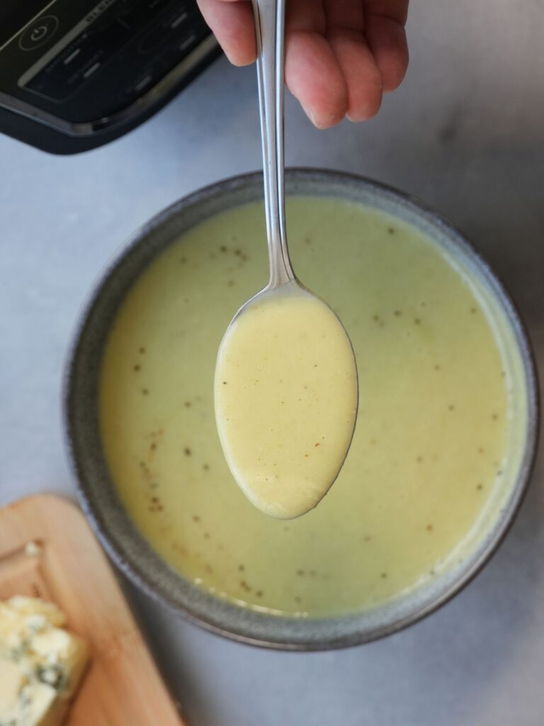 broccoli and stilton soup in a bowl with a spoon of soup held above it. The corner of a ninja soup maker and stilton cheese on a wooden chopping board can just about be seen