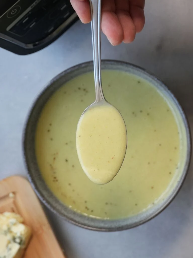 broccoli and stilton soup in a bowl with a spoon of soup held above it. The corner of a ninja soup maker and stilton cheese on a wooden chopping board can just about be seen