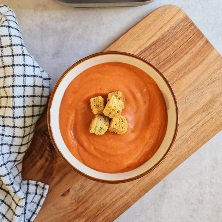 butter bean and lentil soup in a bowl with croutons on top sitting on a wooden chopping board next to a checked tea towel with a little bit of a ninja soup maker showing at the side