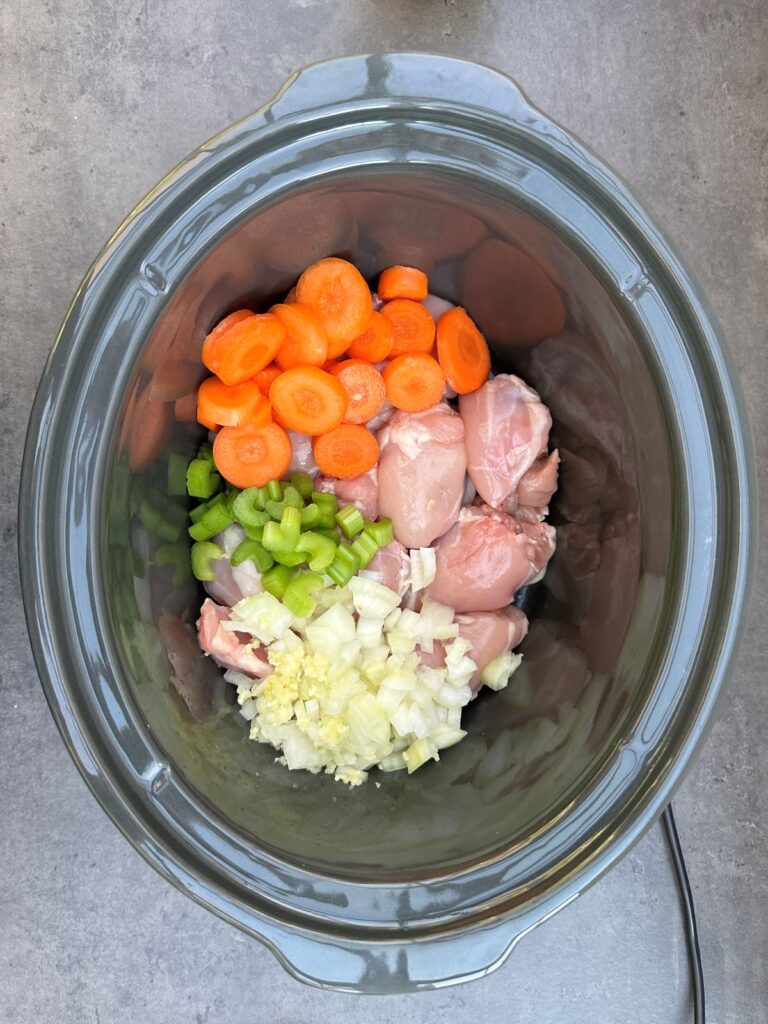 uncooked ingredients for chicken casserole in a slow cooker: boneless chicken thighs, chopped carrots, celery,  onions and crushed garlic