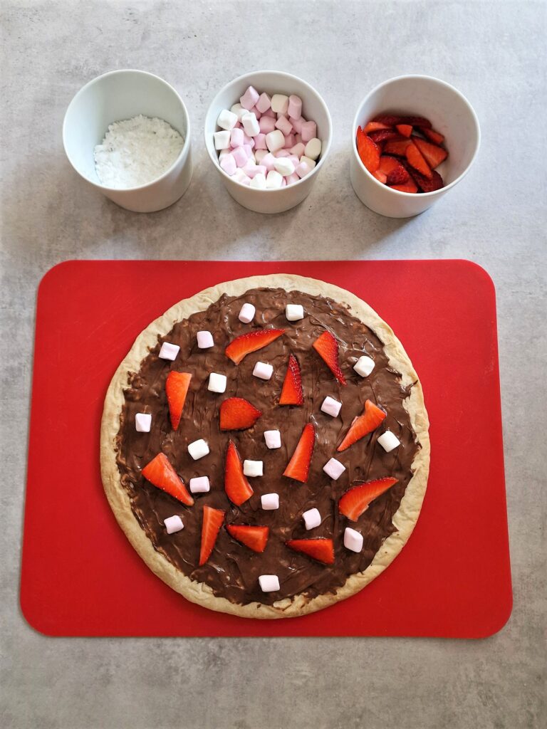 Nutella Chocolate Pizza on red baking mat with 3 bowls of toppings next to it