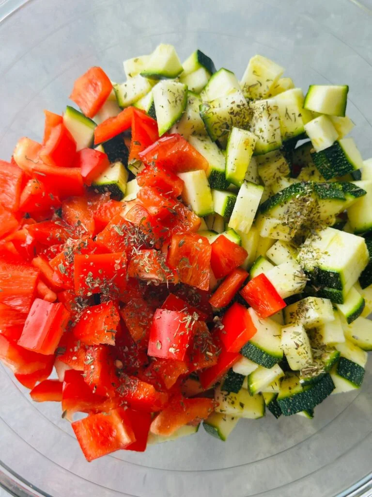 chopped red peppers, courgette, cherry tomatoes, red onion with olive oil and dried mixed herbs on top, in a glass bowl