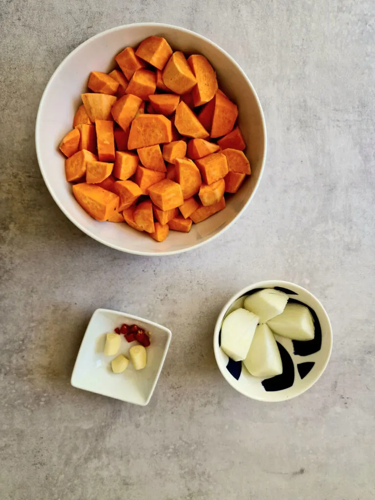 prepared ingredients for soup - peeled and chopped sweet potatoes, quartered raw onion, 3 peeled garlic cloves and a sliced red birds eye chilli