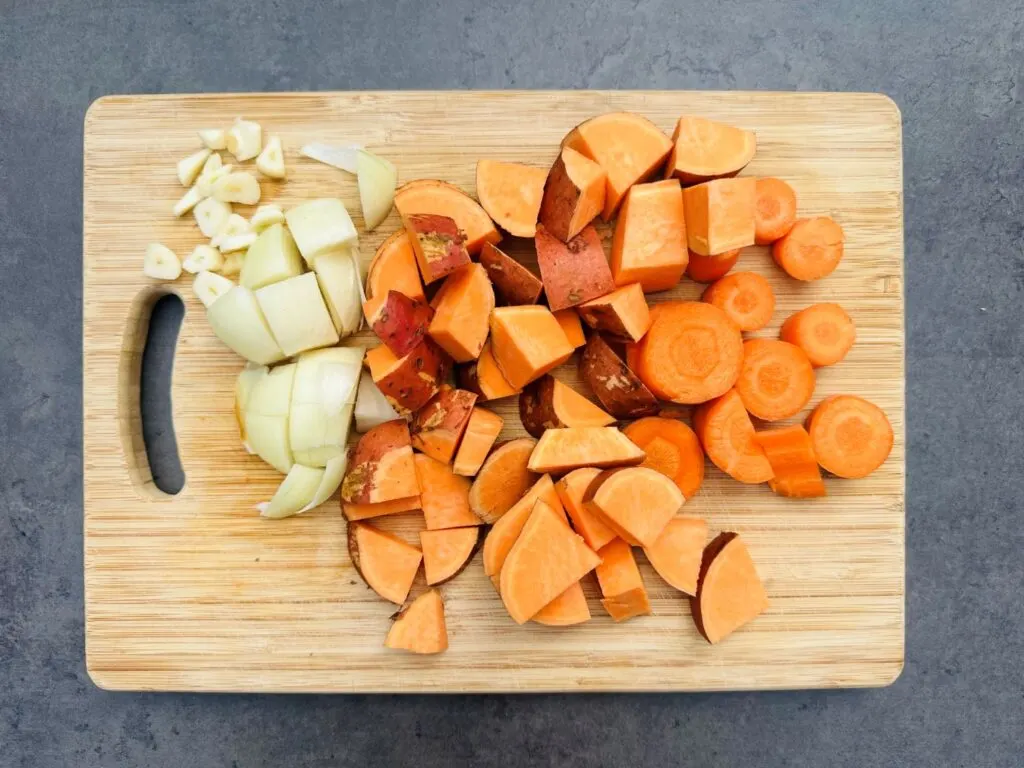 chopped up sweet potato, carrot, onion and garlic on a wooden chopping board