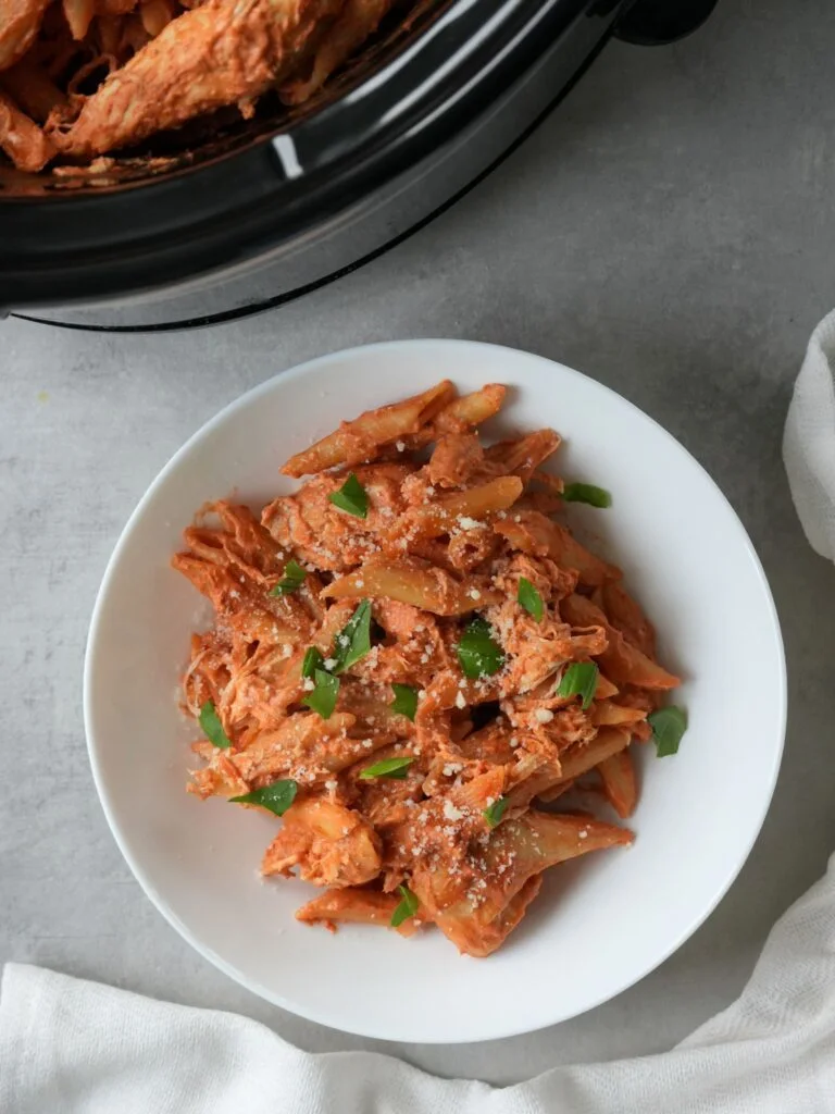 creamy chicken pasta that has been cooked in a slow cooker. Served in a white bowl with fresh torn basil leaves and grated parmesan cheese on top. Part of the slow cooker can been seen in the top left corner.