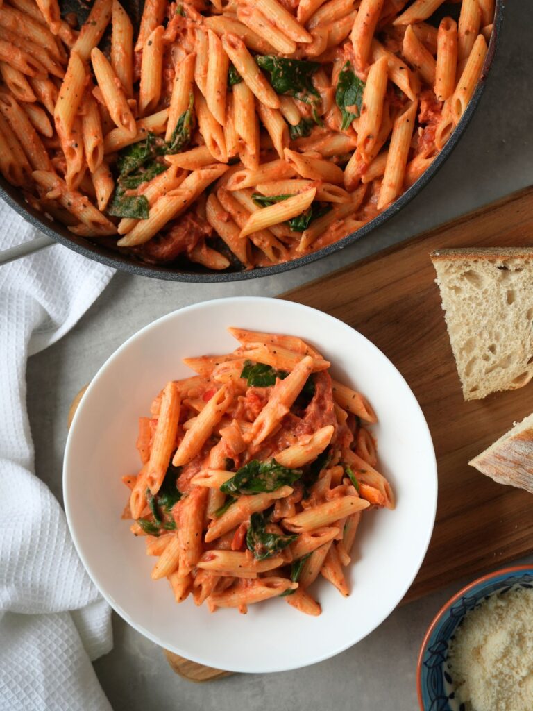 creamy tomato pasta with spinach on a wooden board next to a pan of pasta with slices of bread and grated parmesan cheese in a bowl