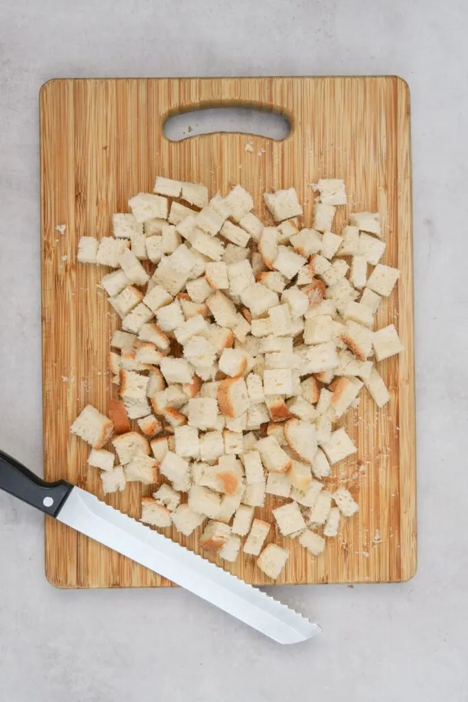 cubed bread for croutons on a chopping board with a shrap bread knife