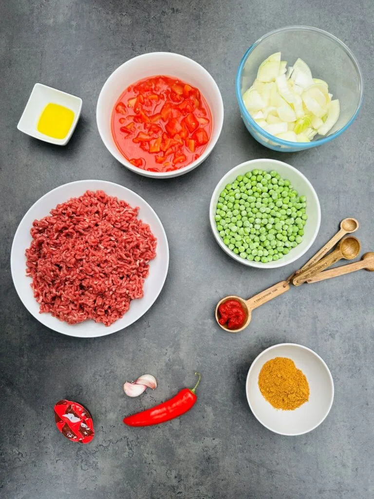 ingredients for cooking curried mince in Ninja Foodi, beef mince, frozen peas, tomato puree, curry powder, two cloves garlic, red chilli, beef stock pot, chopped tomatoes, chopped onion, oil