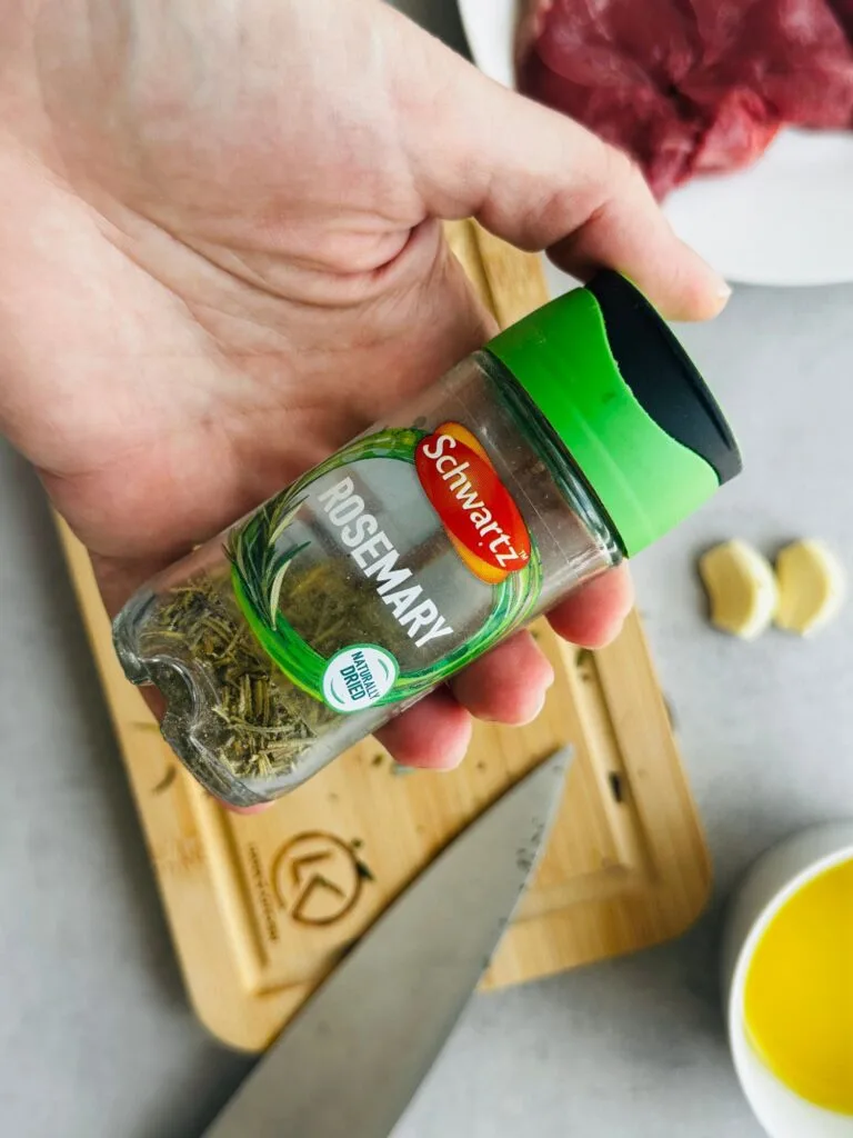 dried rosemary in a Schwartz herb jar being held over a chopping board with two garlic cloves and oil visible in the background