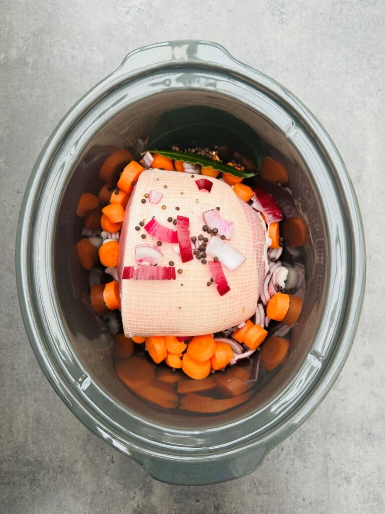 uncooked gammon joint in a slow cooker with chopped carrots, red onion, bay leaf and peppercorns