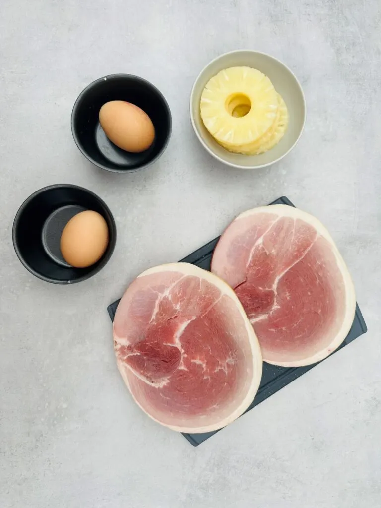 two gammon steaks on chopping board with pineapple slices and eggs in baking tins