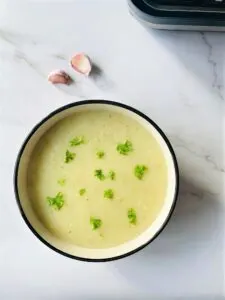 A bowl of potato and garlic soup sprinkled with Basil and a few garlic bulbs to the side