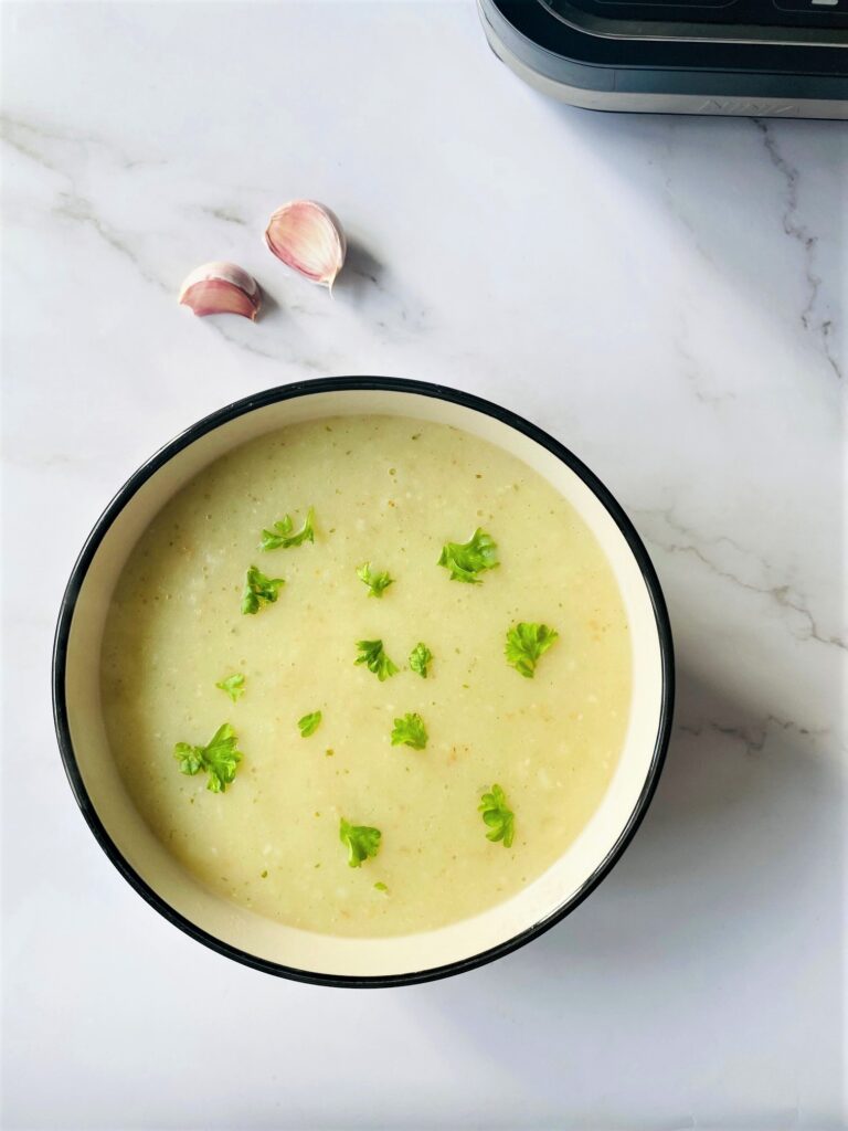 A bowl of potato and garlic soup sprinkled with Basil and a few garlic bulbs to the side 