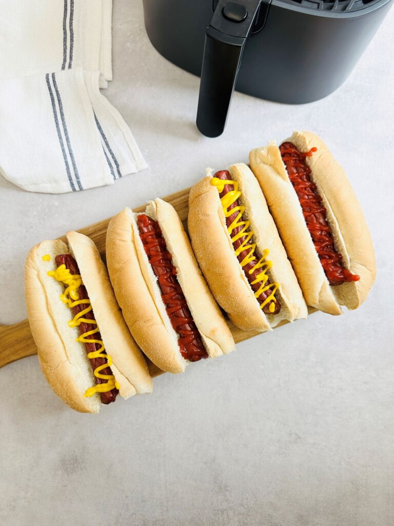 4 hot dogs with mustard and ketchup on a chopping board next to an air fryer
