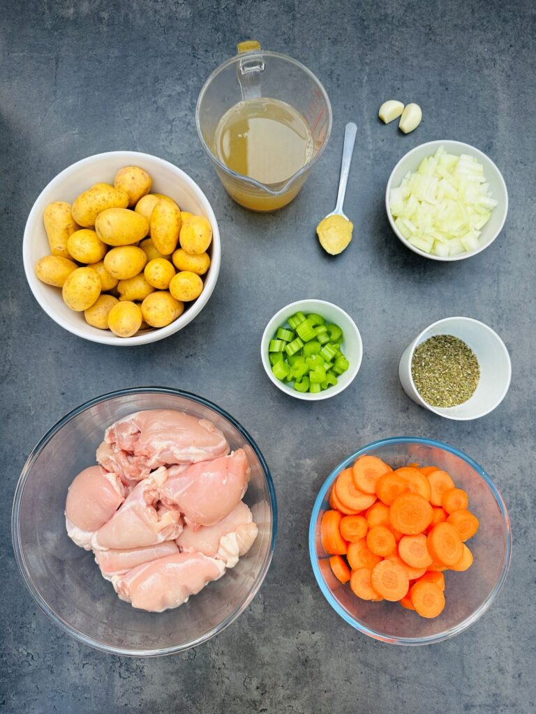 ingredients laid out for cooking chicken casserole in a slow cooker: boneless chicken thighs, chopped carrots, chopped celery, chopped onion, two cloves garlic, 1 tsp mustard, chicken stock, bowl new potatoes