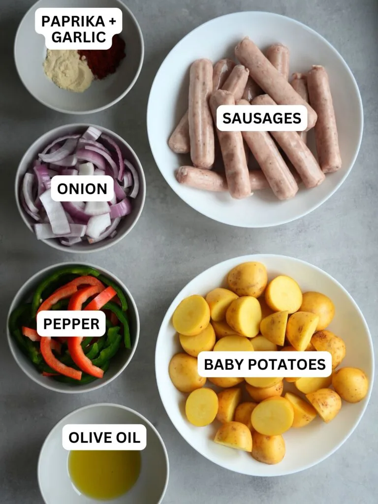 image with ingredients laid out and labelled for air fryer sausage tray bake: paprika and garlic, sausages, onion, pepper, baby potatoes, olive oil