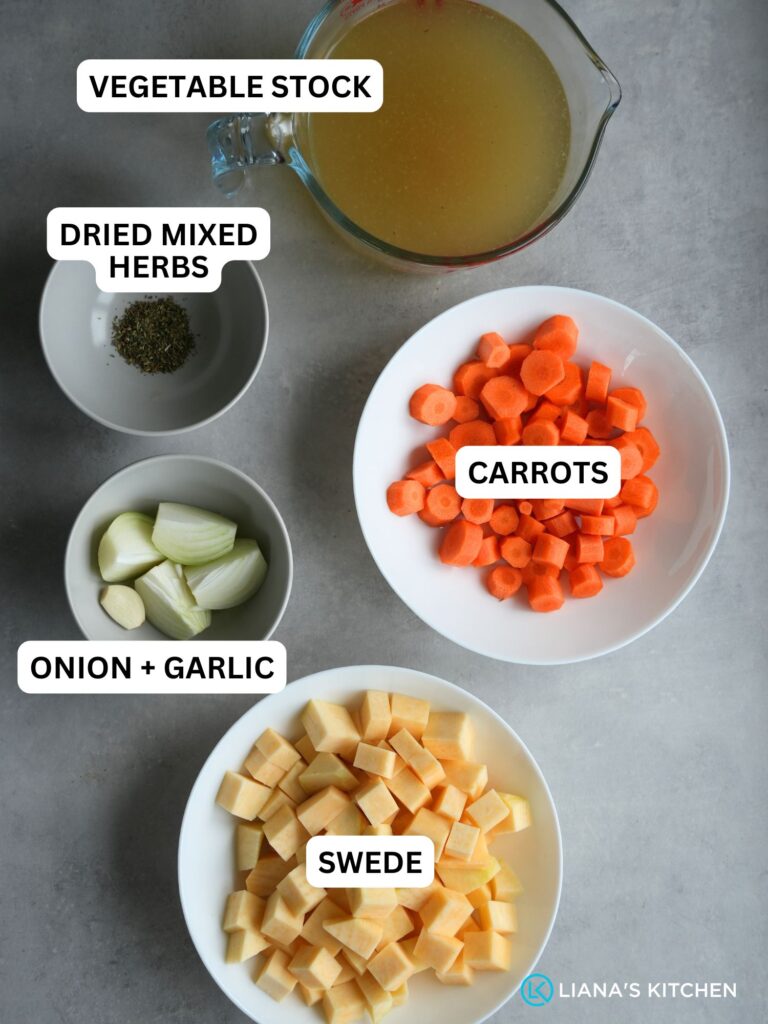 ingredients laid out and labelled for carrot and swede soup in a soup maker: vegetable stock, dried mixed herbs, carrots, onion and garlic, swede