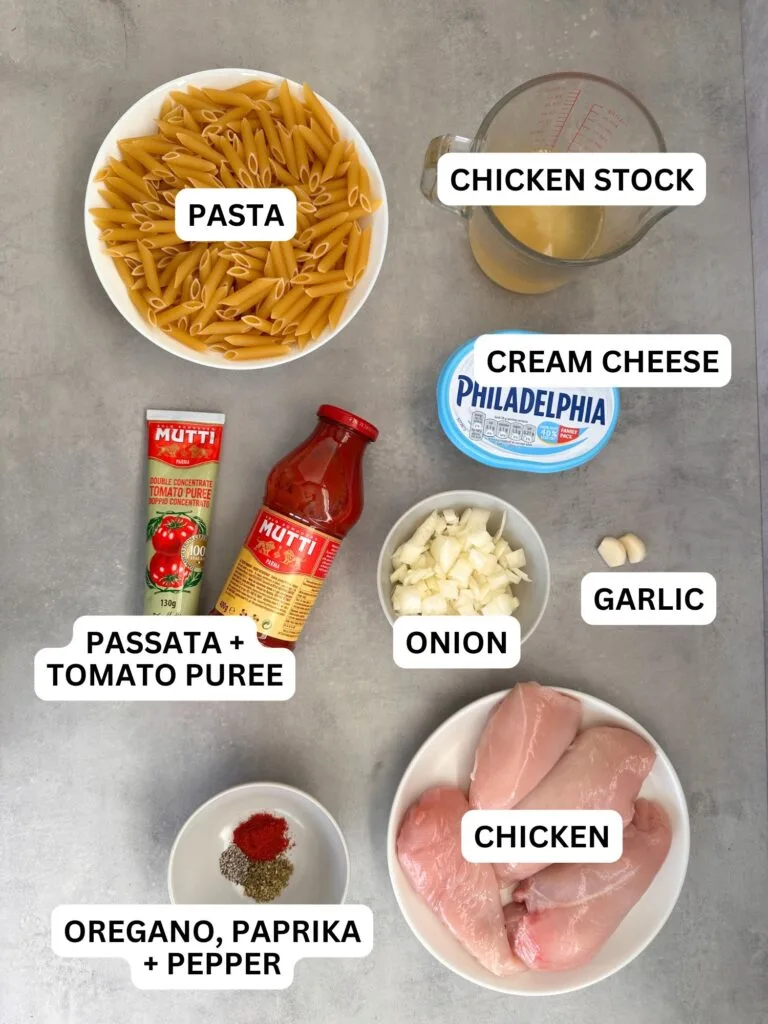 ingredients laid out and labelled for slow cooker creamy chicken pasta: penne pasta, chicken stock, cream cheese, passata and tomato puree, onion, garlic, oregano, paprika, pepper, chicken