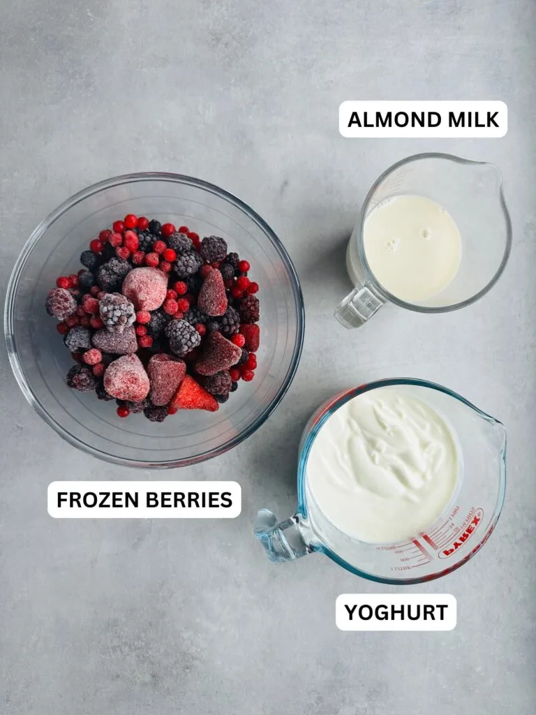 ingredients for frrozen berry smoothie: frozen berries in a glass bowl, almond milk in a jug and yoghurt in a jug